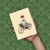 Paquete de imágenes de clipart de mujeres victorianas 8 Una mano sostiene una postal de estilo vintage en la que aparece una mujer con un vestido y un sombrero celestes andando en bicicleta con una cesta. El fondo es un estampado floral verde con textura.