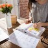 Una mujer sentada ante un escritorio de madera, escribe y subraya notas en una agenda. Sobre el escritorio hay un ordenador portátil, unas gafas, un calendario y un jarrón con rosas naranjas. La luz del sol entra por una ventana cercana.