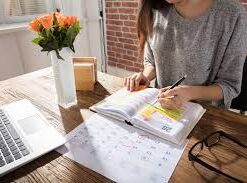 Una mujer sentada ante un escritorio de madera, escribe y subraya notas en una agenda. Sobre el escritorio hay un ordenador portátil, unas gafas, un calendario y un jarrón con rosas naranjas. La luz del sol entra por una ventana cercana.