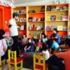 Un hombre se para frente a un pequeño grupo de niños y adultos en una sala colorida, posiblemente una biblioteca o un aula. Parece estar contando historias o actuando. El público, probablemente del nivel "EJERCICIOS GRAMATICALES Y ORTOGRÁFICOS PARA SECUNDARIA", está sentado en el suelo y en sillas, escuchando atentamente, rodeado de estanterías llenas de libros.