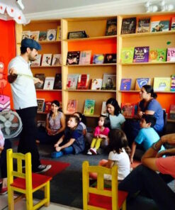 Un hombre se para frente a un pequeño grupo de niños y adultos en una sala colorida, posiblemente una biblioteca o un aula. Parece estar contando historias o actuando. El público, probablemente del nivel "EJERCICIOS GRAMATICALES Y ORTOGRÁFICOS PARA SECUNDARIA", está sentado en el suelo y en sillas, escuchando atentamente, rodeado de estanterías llenas de libros.