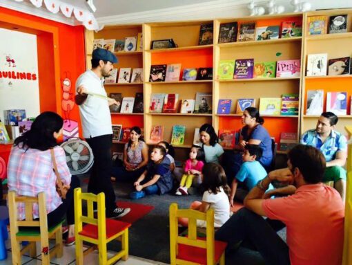 Un hombre se para frente a un pequeño grupo de niños y adultos en una sala colorida, posiblemente una biblioteca o un aula. Parece estar contando historias o actuando. El público, probablemente del nivel "EJERCICIOS GRAMATICALES Y ORTOGRÁFICOS PARA SECUNDARIA", está sentado en el suelo y en sillas, escuchando atentamente, rodeado de estanterías llenas de libros.