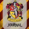 La portada del Diario de Gryffindor o Notas muestra el icónico escudo con un león rampante en dorado y rojo, rodeado por un diseño ornamentado. "Gryffindor" está elegantemente inscrito debajo del escudo, mientras que "Journal o Notas" adorna la parte inferior con una fuente estilizada. El fondo presenta rayas diagonales en amarillo y rojo oscuro.
