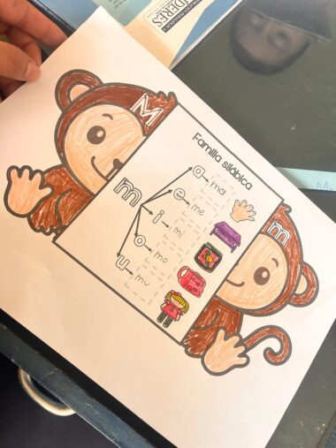 Una mano sostiene una hoja de ejercicios con un mono de dibujos animados y un cuadro titulado "Familia silábica" con las sílabas "ma, me, mi, mo, mu" e imágenes de objetos como una maleta, un calcetín y una magdalena.