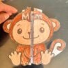 SILABARIO INTERACTIVO COMPLETO CON VOCALES 5 Una mano sostiene una regla vertical de plástico contra el centro de un mono de papel de colores recortado, dividiéndolo en dos mitades simétricas. El mono tiene una