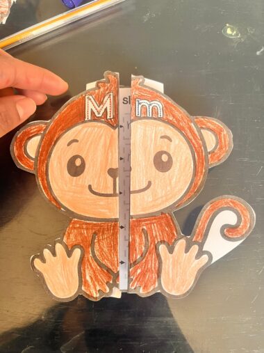 Una mano sostiene una regla vertical de plástico contra el centro de un mono de papel de colores recortado, dividiéndolo en dos mitades simétricas. El mono tiene una "M" y una "m" en la cabeza y está dibujado en marrón, beige y blanco.