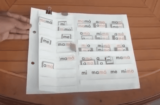Una herramienta de aprendizaje de español hecha a mano para niños, que muestra un papel con solapas etiquetadas con palabras como "mamá", "ama", "mami" y "me" sobre una mesa de madera, diseñada como parte de la serie 22 Cuadernos Dinámicos de Lectura Globalizada. .