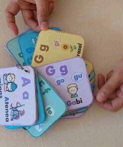 Un primer plano de dos manos hojeando coloridas tarjetas con letras e ilustraciones. Las tarjetas incluyen las letras A, f, G y g con las imágenes y palabras correspondientes. El fondo es una superficie de color beige claro, con LLAVERO REFERENTES SILÁBICOS para fácil referencia.