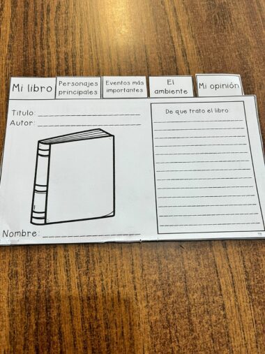 Una hoja de trabajo titulada "Interactivo para reporte de libro" se muestra sobre una superficie de madera. Este reporte de libro interactivo presenta secciones para "Personajes principales", "Eventos más importantes", "El ambiente" y "Mi opinión". Incluye una ilustración del libro vacía, espacios para el título del libro, el autor y un breve resumen.