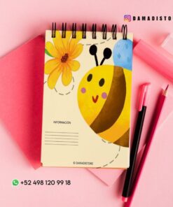 Un cuaderno de espiral con una bonita ilustración de una abeja y una flor en la portada sobre papel rosa, junto a bolígrafos rosas y rojos. Aparecen el usuario de Instagram @damadistore y el número de contacto de WhatsApp.