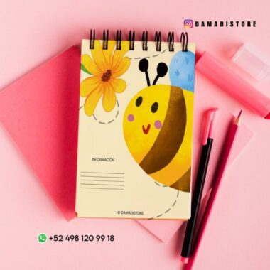 Un cuaderno de espiral con una bonita ilustración de una abeja y una flor en la portada sobre papel rosa, junto a bolígrafos rosas y rojos. Aparecen el usuario de Instagram @damadistore y el número de contacto de WhatsApp.
