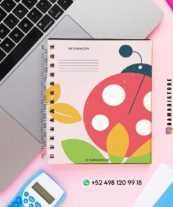Un colorido cuaderno de espiral con un diseño de mariquita está colocado sobre un escritorio junto a un ordenador portátil, una calculadora y un rotulador fluorescente rosa. La cubierta del cuaderno tiene hojas y colores llamativos. Los datos de contacto y las cuentas de redes sociales están visibles.