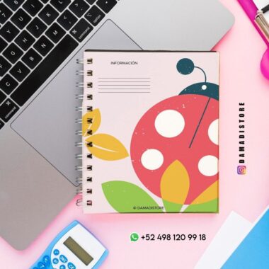 Un colorido cuaderno de espiral con un diseño de mariquita está colocado sobre un escritorio junto a un ordenador portátil, una calculadora y un rotulador fluorescente rosa. La cubierta del cuaderno tiene hojas y colores llamativos. Los datos de contacto y las cuentas de redes sociales están visibles.