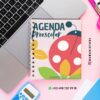 Un cuaderno encuadernado en espiral con un colorido diseño de mariquitas y hojas titulado "Agenda Preescolar" está colocado sobre un escritorio rosa junto a un ordenador portátil, una calculadora azul, un bolígrafo y una tarjeta verde de contacto de WhatsApp.