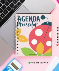 Un cuaderno encuadernado en espiral con un colorido diseño de mariquitas y hojas titulado "Agenda Preescolar" está colocado sobre un escritorio rosa junto a un ordenador portátil, una calculadora azul, un bolígrafo y una tarjeta verde de contacto de WhatsApp.