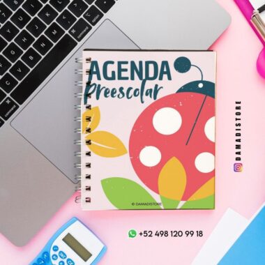 Un cuaderno encuadernado en espiral con un colorido diseño de mariquitas y hojas titulado "Agenda Preescolar" está colocado sobre un escritorio rosa junto a un ordenador portátil, una calculadora azul, un bolígrafo y una tarjeta verde de contacto de WhatsApp.
