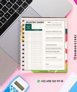 Una hoja de registro diario encuadernada en espiral para profesores está abierta en un ordenador portátil, rodeada de una calculadora, bolígrafos y un teléfono sobre una superficie rosa. La hoja incluye columnas para los nombres de los alumnos, las actividades y diversas observaciones.