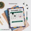 Agenda docente director técnico 2025-2026 emoji 18 Un calendario para septiembre de 2025 está en un cuaderno con una portada azul con temática emoji. Lo rodean objetos como una planta, bolígrafos y clips. Las fechas importantes están marcadas: 8, 15 y 26. En la parte inferior aparecen los datos de contacto.