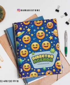 Un colorido cuaderno de espiral titulado "AGENDA DEL DIRECTOR" con emojis de sonrisas y estrellas en la portada, colocado encima de otros cuadernos, junto a un bolígrafo, un lápiz, clips y una pequeña maceta sobre una superficie blanca.