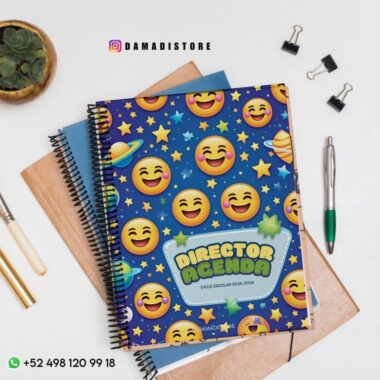Un colorido cuaderno de espiral titulado "AGENDA DEL DIRECTOR" con emojis de sonrisas y estrellas en la portada, colocado encima de otros cuadernos, junto a un bolígrafo, un lápiz, clips y una pequeña maceta sobre una superficie blanca.