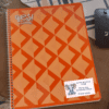 Etiquetas para cuadernos, libros y lápices de Elefantes 3 Sobre un escritorio hay un cuaderno de espiral naranja con un patrón geométrico. Una pegatina blanca en la portada dice