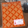 Etiquetas para cuadernos, libros y lápices 3 Sobre un escritorio hay un cuaderno naranja con un patrón geométrico, junto a un ratón y un objeto con rayas azules. Una etiqueta con su nombre en el cuaderno dice