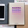 Un cuaderno de espiral con la palabra "MAESTRA" escrita cuatro veces en diferentes tonos de morado y rosa. El cuaderno está sobre un escritorio junto a un smartphone. En la imagen se ven los datos de contacto y la dirección de las redes sociales.