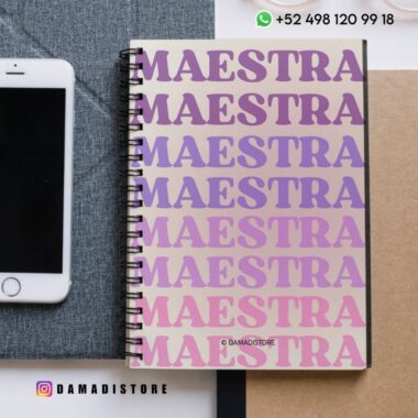 Un cuaderno de espiral con la palabra "MAESTRA" repetida en grandes letras moradas y rosas en la portada, junto a un smartphone, un lápiz y un bloc de notas. En la imagen aparecen los datos de contacto de la tienda y su cuenta de Instagram.