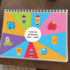 Un colorido cuaderno con la lista de asistencia para 2024-2025, abierto sobre una mesa. la portada presenta lindos íconos de dibujos animados como un oso, una manzana, un helado y otros, divididos por secciones multicolores.