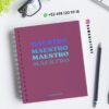 Un cuaderno de espiral con "MAESTRO" escrito cuatro veces en diferentes tonos de azul en la portada, colocado sobre un escritorio en el que se muestran gafas, un bolígrafo, una planta y la información de contacto de DAMADISTORE.