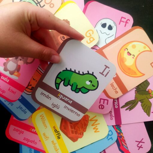 Una mano sostiene una tarjeta con la letra "I" y una iguana de dibujos animados, junto con las palabras en español "iguana, imán, iglú, invierno". Las Tarjetas del abecedario (Lectura de palabras) circundantes presentan varias letras, ilustraciones coloridas y palabras en español.