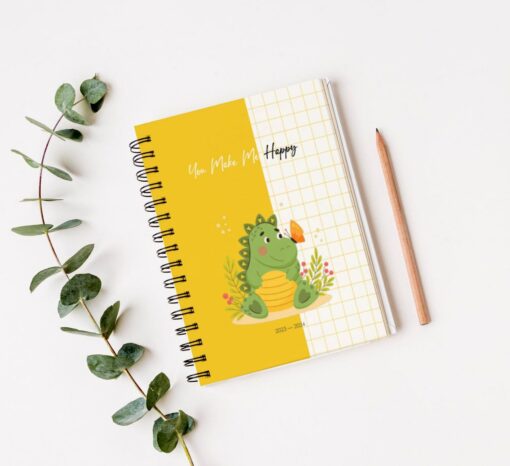 Un Cuaderno de estudio - Dinosaurio (100 hojas) encuadernado en espiral y con tapa amarilla que presenta una caricatura de un dinosaurio verde sentado entre plantas, sosteniendo una mariposa. El texto "You Make Me Happy" está impreso encima del dinosaurio. Un lápiz y una ramita de hojas se colocan al lado del Cuaderno de estudio - Dinosaurio (100 hojas).