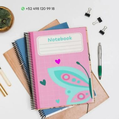 Un Cuaderno de raya Mariposa PDF con una cubierta rosa con un diseño de Mariposa se encuentra encima de otros artículos de papelería, incluido un cuaderno azul y uno marrón. Hay un bolígrafo, dos clips para carpetas y una pequeña planta en una maceta cerca. Se ve un número de contacto de WhatsApp.