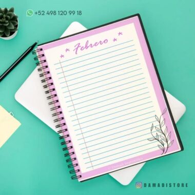 Un cuaderno de raya encuadernado en espiral abierto en una página titulada "Febrero" con bordes, líneas y una ilustración de flores en la esquina inferior derecha. Se muestra un número de teléfono en la parte superior izquierda y hay una planta suculenta, un Cuaderno de raya con diseño de rana cerrado y el identificador "@DAMADISTORE" cerca.