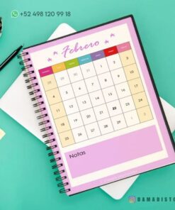 Un cuaderno de raya con diseño de rana encuadernado en espiral descansa sobre una superficie verde azulado que muestra un colorido calendario de febrero con fechas en una cuadrícula. La información de contacto y un identificador de redes sociales son visibles. Al fondo se ve una planta suculenta y un teclado parcialmente visible.