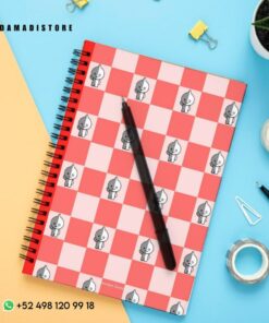 Sobre un escritorio hay un cuaderno de espiral con una cubierta a cuadros rojos y blancos con pequeños personajes de dibujos animados BT21. Un bolígrafo negro descansa sobre el Cuaderno BT21 VAN. Los elementos circundantes incluyen cinta adhesiva, una planta en maceta, una carpeta amarilla, clips y una perforadora pequeña.