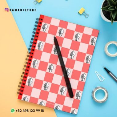 Sobre un escritorio hay un cuaderno de espiral con una cubierta a cuadros rojos y blancos con pequeños personajes de dibujos animados BT21. Un bolígrafo negro descansa sobre el Cuaderno BT21 VAN. Los elementos circundantes incluyen cinta adhesiva, una planta en maceta, una carpeta amarilla, clips y una perforadora pequeña.
