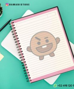 Un Cuaderno BT21 Shooky con cubierta rosa y páginas rayadas se encuentra encima de una tableta blanca. Un personaje estilizado de dibujos animados Shooky de BT21 está superpuesto en el cuaderno. A su alrededor se ven un bolígrafo negro, una pequeña planta, una nota adhesiva e información de contacto.