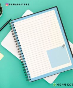 Un Cuaderno BT21 Shooky encuadernado en espiral está abierto y muestra páginas rayadas con una sección para notas. Cerca hay un bolígrafo negro, tarjetas pequeñas con Shooky de BT21 y una planta suculenta. Al fondo hay una tableta y un teléfono. La imagen incluye los datos de contacto de Instagram y WhatsApp de Dama Di Store.