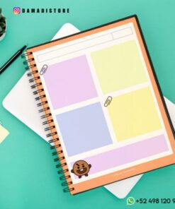 Se coloca un Cuaderno BT21 Shooky con secciones codificadas por colores sobre una superficie verde azulado. Un emoji con una cara marrón sonriente, que se parece a Shooky de BT21, adorna la sección inferior. Cerca hay un bolígrafo, una nota adhesiva y una pequeña planta suculenta. El cuaderno tiene la marca 