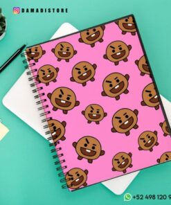 Un Cuaderno BT21 Shooky se coloca encima de un cuaderno blanco. Cerca hay un bolígrafo negro, una planta suculenta en una maceta y una pequeña tarjeta amarilla. Las superposiciones de texto incluyen información de la tienda y de contacto.
