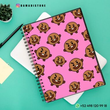 Un Cuaderno BT21 Shooky se coloca encima de un cuaderno blanco. Cerca hay un bolígrafo negro, una planta suculenta en una maceta y una pequeña tarjeta amarilla. Las superposiciones de texto incluyen información de la tienda y de contacto.