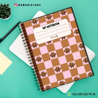 Un cuaderno de espiral sobre un escritorio con una cubierta de cuadros rosas y marrones con pequeñas ilustraciones de osos. La etiqueta de la portada dice "MI CUADERNO" con "Encantadora" debajo. Cerca hay una tableta blanca y una pequeña planta en una maceta. Los datos de contacto son visibles y está al lado de un Cuaderno BT21 Shooky.
