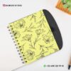 Cuaderno Descripción de Mis Libros leídos 17 Un *Cuaderno Descripción de Mis Libros leídos* encuadernado en espiral abierto que muestra una cubierta amarilla con diseños florales negros. El cuaderno está colocado sobre una superficie blanca con un objeto de madera parcialmente visible en la parte superior derecha y una pequeña planta suculenta verde en la parte superior derecha. El texto incluye el identificador de Instagram @DAMADISTORE y el número de contacto de WhatsApp +52 498 120 99 18.