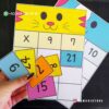Tablero de las multiplicaciones (carta) 6 Una mano sostiene coloridas tarjetas numéricas mientras las organiza en una cuadrícula con temática de gatos de color amarillo brillante y azul. Cada tarjeta tiene números diferentes del Tablero de multiplicaciones (carta multimedia), incluidos el 7 y el 21. La información de contacto de WhatsApp y el nombre de la tienda se muestran en la imagen de la carta multimedia.