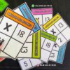 Una mano que sostiene coloridas tarjetas de multiplicación que muestran ecuaciones como 3x11 y 6x18. Algunas tarjetas, centradas en *Mis multiplicaciones del 11-20*, están dispuestas sobre una superficie negra. El texto en la parte superior muestra un número de WhatsApp, mientras que 
