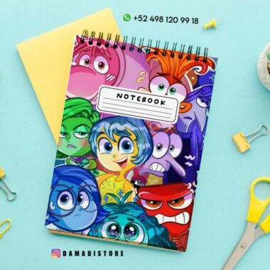 Un colorido cuaderno con encuadernación en espiral con varios personajes animados en la portada. El fondo es una superficie azul claro con tijeras amarillas dispersas, un pequeño cactus en una maceta, clips y un sobre amarillo. Los datos de contacto están en la parte superior.