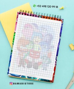 Un cuaderno de espiral cuadriculado con una colorida cubierta animada con varios personajes de dibujos animados se asienta sobre una superficie azul claro. Cerca hay un par de tijeras amarillas, clips metálicos, una carpeta amarilla, una planta y un identificador de Instagram 