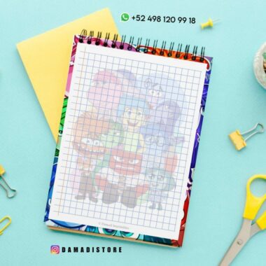 Un cuaderno de espiral cuadriculado con una colorida cubierta animada con varios personajes de dibujos animados se asienta sobre una superficie azul claro. Cerca hay un par de tijeras amarillas, clips metálicos, una carpeta amarilla, una planta y un identificador de Instagram "@DAMADISTORE" con un número de teléfono.