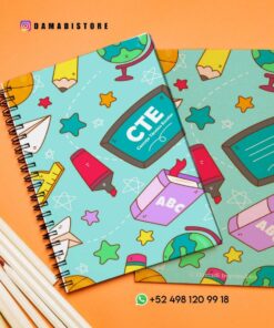 Sobre un escritorio con lápices se exponen dos cuadernos ilustrados. Las portadas presentan coloridos diseños con temas escolares, que incluyen globos terráqueos, libros, lápices e íconos relacionados con la educación. El identificador @DamaDiStore y un número de teléfono son visibles.